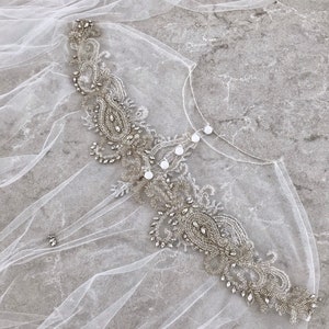 May include: A delicate white tulle bridal veil with a beaded embellishment featuring a paisley design and clear rhinestones.