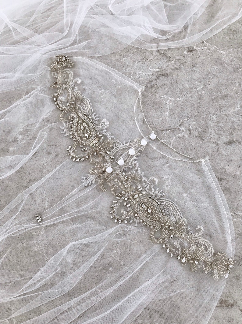 May include: A delicate white tulle bridal veil with a beaded embellishment featuring a paisley design and clear rhinestones.
