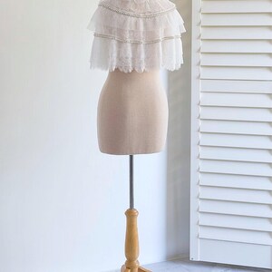 May include: A white lace bridal cape with pearl embellishments, displayed on a wooden mannequin stand.