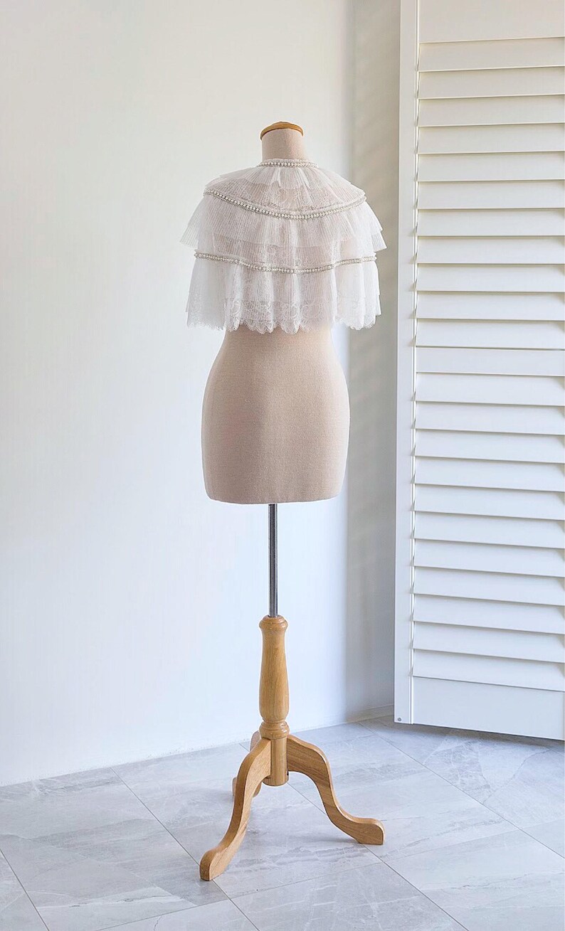 May include: A white lace bridal cape with pearl embellishments, displayed on a wooden mannequin stand.
