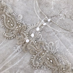 May include: A white tulle fabric with a beaded trim. The trim features a paisley design with clear and silver beads.