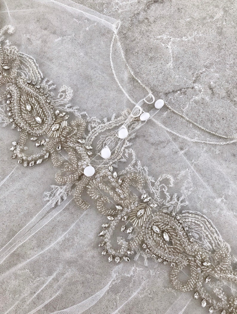 May include: A white tulle fabric with a beaded trim. The trim features a paisley design with clear and silver beads.