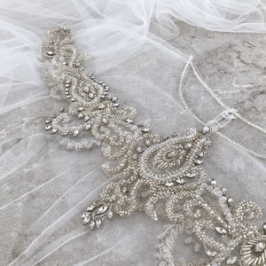 May include: A delicate white tulle fabric with a beaded and sequined embellishment. The embellishment features a paisley design with clear and silver beads.