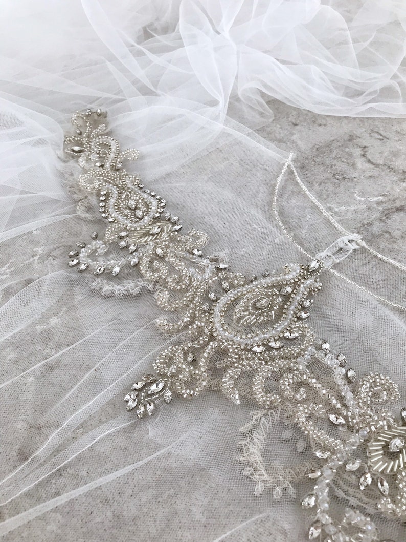 May include: A delicate white tulle fabric with a beaded and sequined embellishment. The embellishment features a paisley design with clear and silver beads.