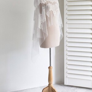 May include: A white lace bridal shawl with a high neckline and a sheer overlay. The shawl is displayed on a wooden mannequin stand.