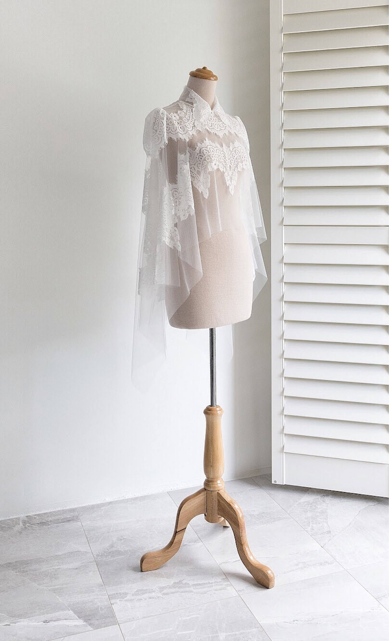 May include: A white lace bridal shawl with a high neckline and a sheer overlay. The shawl is displayed on a wooden mannequin stand.