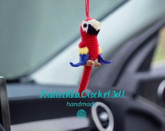 Può includere: Un ciondolo per auto a forma di pappagallo all'uncinetto fatto a mano. Il pappagallo è principalmente rosso con dettagli gialli, blu e bianchi. Il ciondolo è appeso a un filo rosso. Il testo "Kralechka Crochet Doll handmade" è visibile.