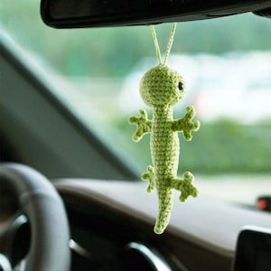 Puede incluir: Un gecko de crochet verde colgando de un espejo retrovisor en un coche. El gecko tiene ojos negros y una cola larga.