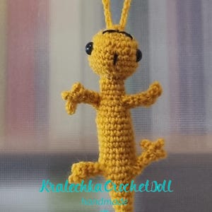May include: A yellow crocheted gecko with black eyes and a black mouth. The gecko is hanging from a string and has the text "Kralecka Crochet Doll handmade" below it.