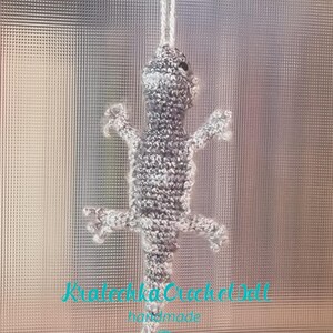 Może przedstawiać: Szara szydełkowa jaszczurka wisząca na białym sznurku. Jaszczurka ma białą plamkę na głowie. Widoczny jest tekst "Kraleczka Crochet Doll handmade" pod jaszczurką.