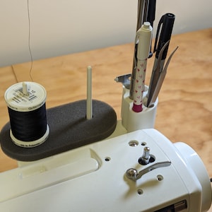 May include: A white sewing machine with a black thread spool, a white bobbin holder, and a black foam pad. A white plastic organizer with a pink base holds pens, tweezers, and a small tool.