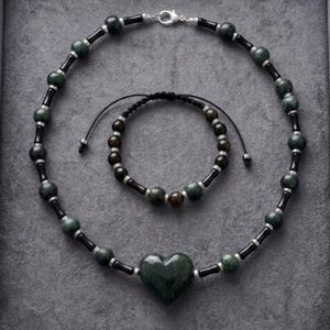 Puede incluir: Un conjunto de collar y pulsera con cuentas de color verde oscuro y negro y separadores plateados. El collar incluye un colgante central en forma de corazón verde oscuro, mientras que la pulsera ajustable tiene cuentas en un cordón negro.