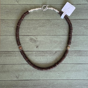 May include: A brown wooden beaded necklace with silver accents and a silver clasp. The necklace has two small gold beads on either side of the clasp. The necklace is on a light green wooden background. The tag attached to the necklace reads "Camp 61 Jewelry Handmade in Albuquerque, NM."