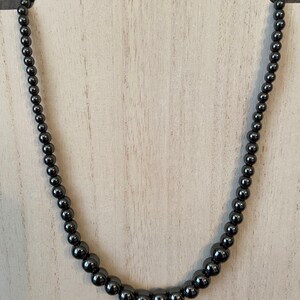 May include: A black beaded necklace with round beads. The necklace is displayed on a white wooden surface.