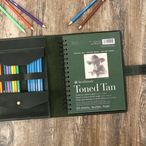 May include: An open, dark green leather art journal with a spiral-bound Strathmore Toned Tan sketch pad. The journal has a pencil holder filled with colored pencils. Several colored pencils are scattered around the journal.