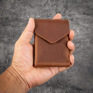 May include: A brown leather wallet featuring a smooth matte finish, a rectangular shape, and a pointed flap closure. The wallet is constructed with visible stitching along the edges, creating a clean and simple design for holding cards or cash.