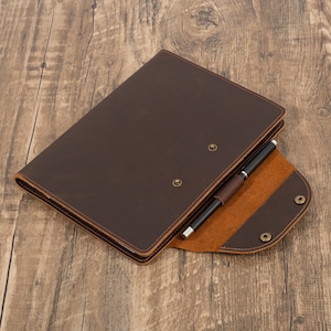 May include: A brown leather notebook with a pen holder and a flap closure. The notebook is open to reveal a tan leather interior.