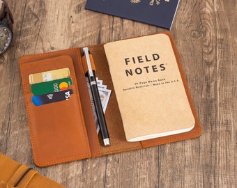 May include: A brown leather notebook cover with a pen, credit cards, and cash inside. The notebook cover has a pocket for cards and a slot for a pen. The notebook is a Field Notes 48-Page Memo Book. The cover is open and the notebook is visible. The notebook is made in the USA.