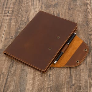 May include: A brown leather portfolio with a pen holder and a black pen. The portfolio is open, revealing a tan interior. The cover is secured with snap closures. The item is on a wooden surface.