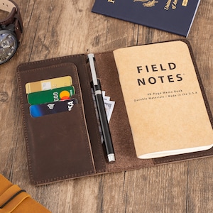 May include: A brown leather notebook cover with a pen holder, card slots, and a Field Notes memo book inside. The cover is open and the memo book is visible. The memo book is brown and has the words "Field Notes" printed on the front.