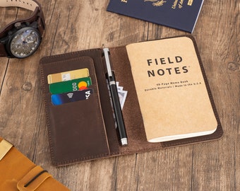 May include: A brown leather notebook cover with a pen holder, card slots, and a Field Notes memo book inside. The cover is open and the memo book is visible. The memo book is brown and has the words "Field Notes" printed on the front.