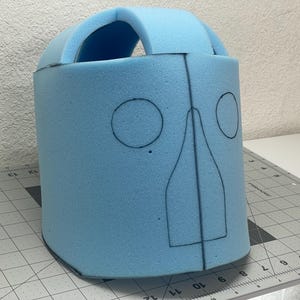 Peut inclure: Une base de casque en mousse bleu clair avec une partie supérieure incurvée et un panneau avant marqué de contours à l'encre noire représentant deux trous oculaires circulaires et une pièce nasale centrale, posée sur un tapis de découpe gris.