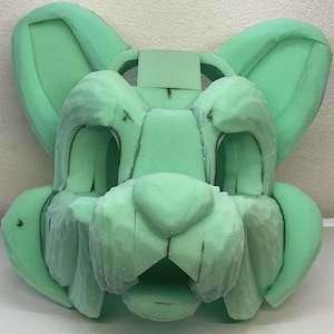 May include: A green foam base for a furry costume head. The base has been sculpted to resemble a cat's face with ears, a nose, and a mouth.