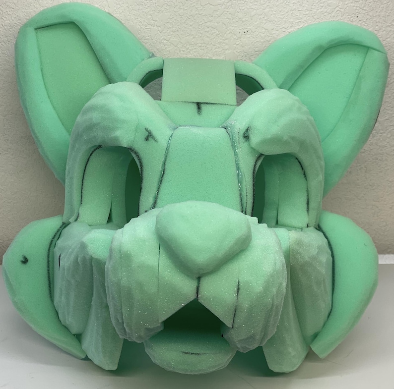 May include: A green foam base for a furry costume head. The base has been sculpted to resemble a cat's face with ears, a nose, and a mouth.