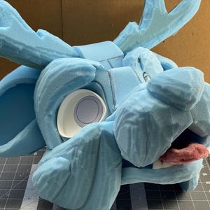 May include: A light blue foam moose head with large antlers, a white eye socket, and a pink tongue. The head is in the process of being constructed, with visible seams and unfinished edges. The head is on a gray cutting mat.