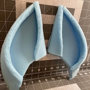 Puede incluir: Dos formas de orejas de espuma azul claro, posiblemente para un disfraz o un proyecto de manualidades. La espuma es de color liso con una superficie ligeramente texturizada. Las orejas están cortadas y listas para el montaje. El fondo es una esterilla de corte con medidas.