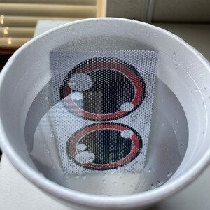 May include: Two red and black cartoon eyes printed on a white mesh sheet submerged in a white styrofoam cup filled with water.