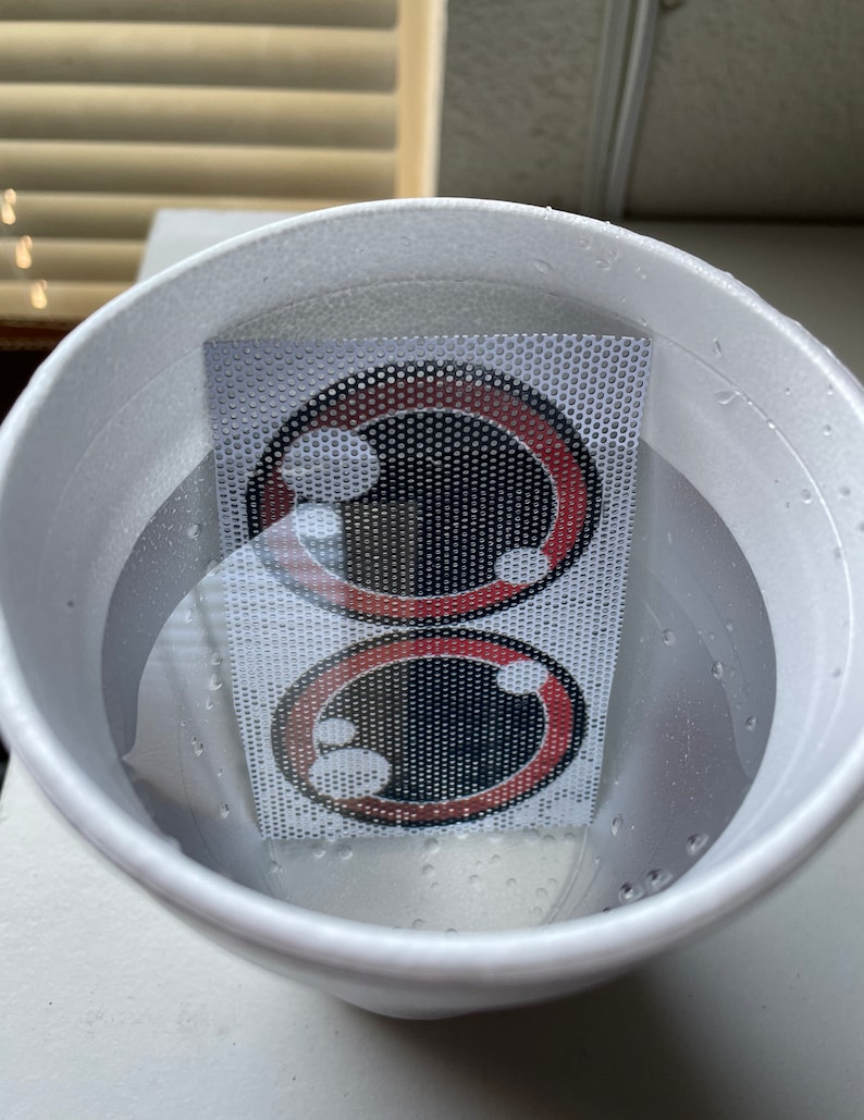 May include: Two red and black cartoon eyes printed on a white mesh sheet submerged in a white styrofoam cup filled with water.