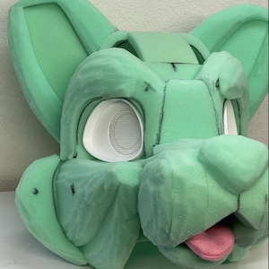 May include: A green foam base for a furry costume head. The base has two ears, a snout, and a tongue. The eyes are cut out and have white foam circles with mesh behind them.
