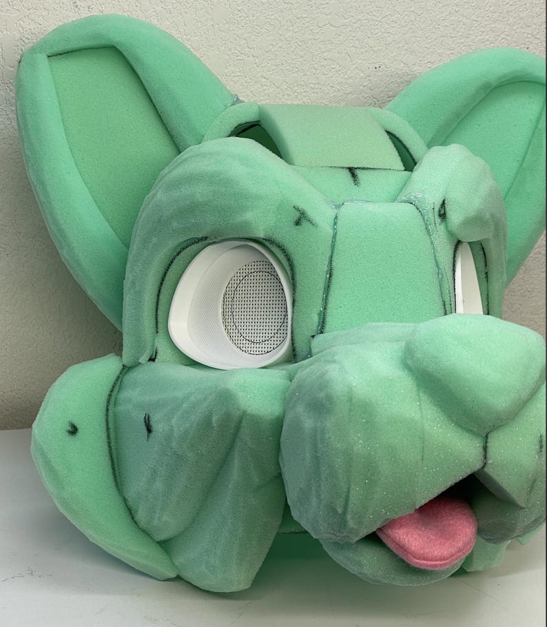May include: A green foam base for a furry costume head. The base has two ears, a snout, and a tongue. The eyes are cut out and have white foam circles with mesh behind them.