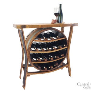 May include: A rustic wine rack made from a repurposed wooden barrel. The rack features a circular wine storage area with multiple shelves, holding several wine bottles. A wooden tabletop holds a wine glass, a bottle of wine, and grapes. The rack is supported by curved wooden legs.