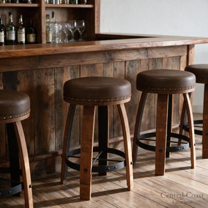 May include: Three bar stools with round brown leather seats, brass nailhead trim, and curved wooden legs reinforced by a black metal circular footrest. The stools are positioned in front of a rustic wooden bar counter with a dark wood finish.