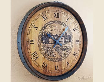 May include: A wooden clock with Roman numerals and a black metal frame. The clock face features a black and white engraved image of a house and vineyard. The clock is 12 inches in diameter.