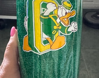 May include: A green glitter tumbler featuring a large yellow and green letter O logo with a cartoon duck mascot wearing a green and yellow cap with the word OREGON printed on it. The tumbler has a clear plastic lid and a smooth, shiny finish.