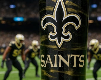 May include: A cylindrical tumbler featuring a black and gold swirled pattern with the New Orleans Saints fleur-de-lis logo and the word SAINTS printed in gold lettering on the front surface.