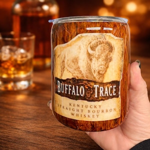 Puede incluir: Un vaso cilíndrico con patrón de veta de madera que presenta un gráfico de un búfalo y el texto Buffalo Trace Kentucky Straight Bourbon Whiskey. La taza tiene una tapa transparente y un acabado liso, mostrando un logotipo de marca detallado.
