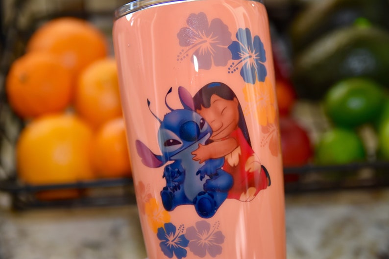 May include: A pink stainless steel tumbler with a graphic of Stitch and Lilo from the Disney movie Lilo & Stitch. The graphic shows Stitch hugging Lilo, who is wearing a red and yellow dress. The tumbler is decorated with pink, blue, and yellow hibiscus flowers.