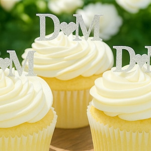 Könnte beinhalten: Drei Cupcakes mit weißem Zuckerguss, jeweils gekrönt mit einem silbernen Glitzer-Kuchentopper, der die Buchstaben D und M zeigt, getrennt durch ein Herzsymbol. Die Topper bestehen aus einem dünnen, flachen Material und stehen aufrecht.