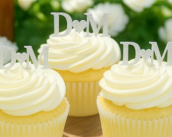 Pode incluir: Três cupcakes com cobertura branca, cada um coberto com um topo de bolo de glitter prateado apresentando as letras D e M separadas por um símbolo de coração. Os topos são feitos de um material fino e plano e ficam na vertical.