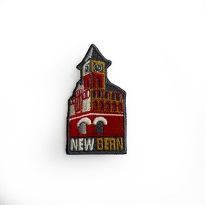 May include: Embroidered patch featuring a red brick building with a clock tower and the text "NEW BERN" in black and gold lettering.