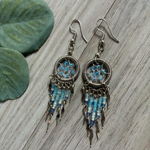 May include: Silver-toned dangle earrings with a dreamcatcher design. Each earring features a circular frame with a web of blue beads, dangling strands of blue and white beads, and silver metal accents. The earrings are on a wooden surface.