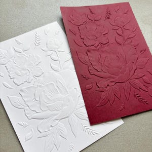 May include: Two rectangular cards, one white and one deep red, each featuring a raised, embossed floral design. The pattern on both cards displays large, detailed peony-like flowers surrounded by smaller leaves and delicate sprigs.