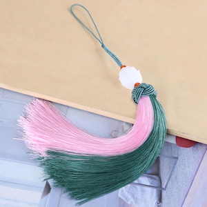 May include: A decorative tassel ornament featuring a long loop of teal cord, a white carved stone bead, and two small orange accent beads. The tassel itself is composed of a dense, layered fringe in alternating shades of soft pink and deep teal.