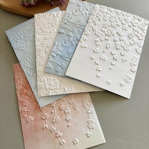 May include: A set of four rectangular cards featuring embossed floral patterns. The cards display various designs including trailing blossoms, intricate vines, and large flower heads in shades of white, pale blue, and a soft peach gradient.