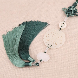 May include: A decorative hanging ornament featuring a dark green silk tassel, a carved off-white circular jade-like pendant with intricate patterns, a small translucent bead, and a dark green cord tied in a complex knot at the top end.