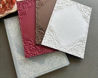 May include: A collection of four rectangular embossing folders featuring intricate, raised border patterns. The set includes one translucent plastic folder, one deep red cardstock, one dark brown cardstock, and one white cardstock.